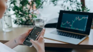 Persona analizando gráficos financieros en su teléfono móvil y laptop para invertir en acciones tecnológicas con bajo capital